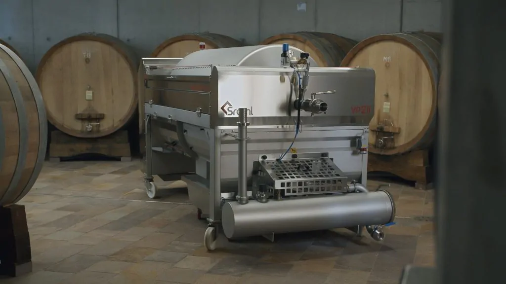 Stainless steel industrial wine processing machine on wheels in a winery cellar with wooden barrels in the background.