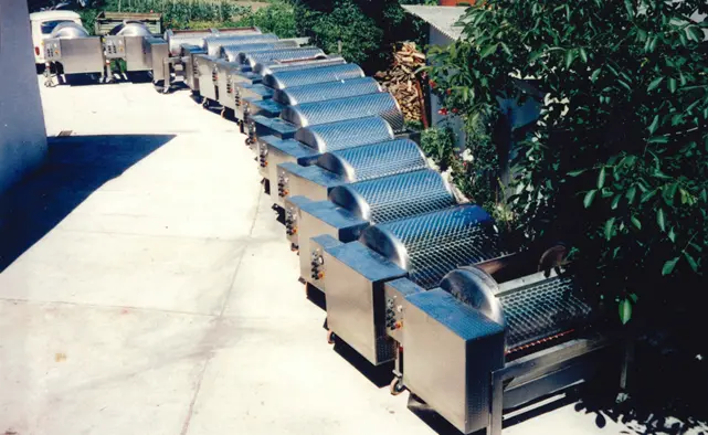 Row of stainless steel outdoor solar dryers with curved vent covers on a concrete patio, greenery in the background.