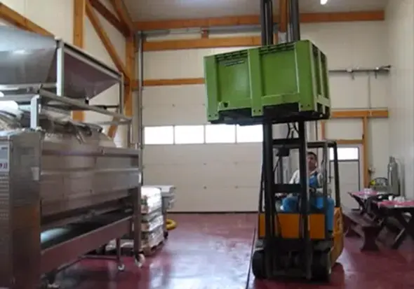 Forklift operator moves a large green crate in a warehouse with metal shelving and a red floor.