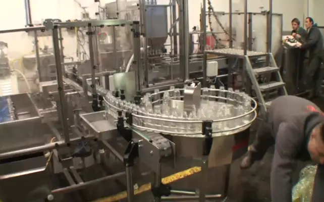 Industrial bottling line with a circular glass bottle carousel and stainless steel machinery in a factory setting; workers in the background.