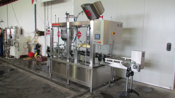 Industrial stainless steel packaging machine with a hopper on top, conveyor belt, and control panel in a warehouse setting.