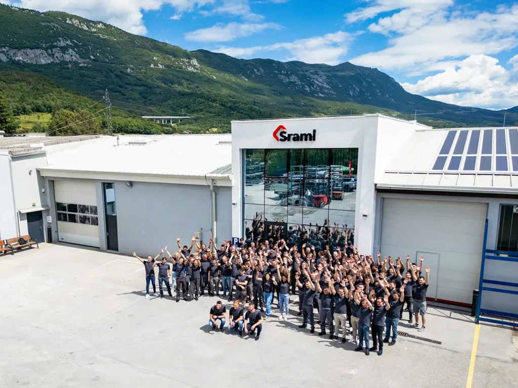 Group of employees in dark shirts posing outside the Sraml building with mountains in the background, arms raised in celebration.