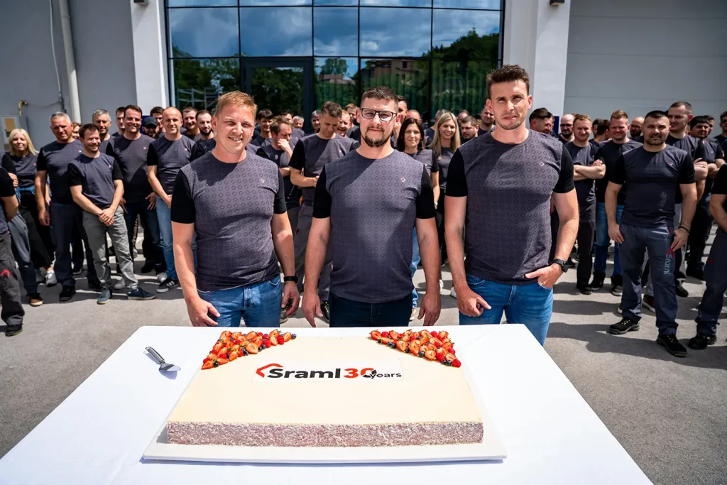 Three men in matching gray shirts stand at a table with a large cake, strawberries on top, while coworkers gather behind them outside a modern building. The cake bears a "Sraml 30 years" logo and celebration theme shows.