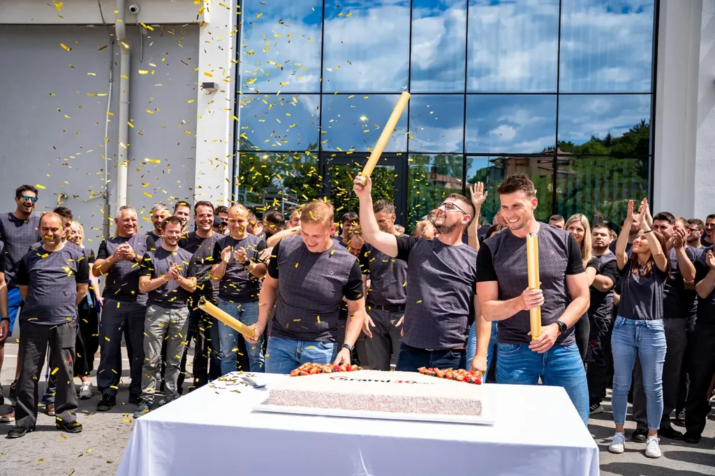 Group of coworkers in dark gray shirts cheer as yellow confetti falls around a table with a large cake outside a glass-fronted building, one man raising a celebratory tube in the air.