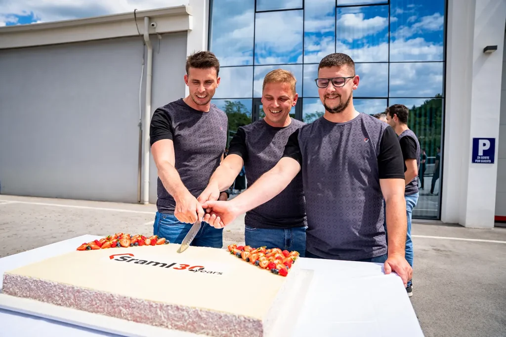 Three men in matching dark shirts slice a large celebratory cake outdoors in front of a glass-front building.