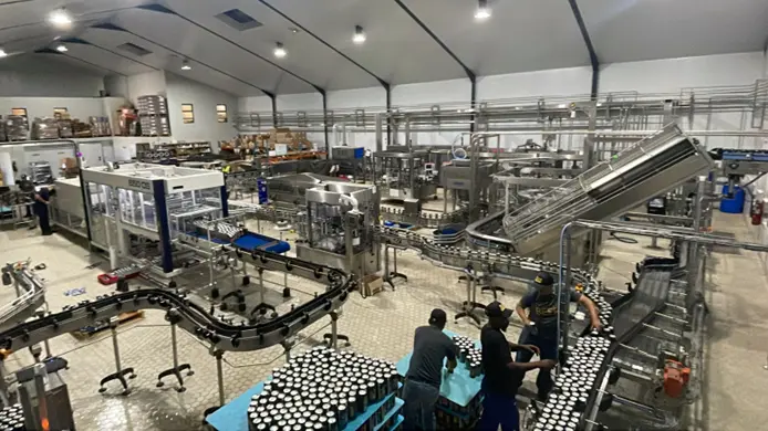 Inside a large beverage bottling plant with stainless equipment, conveyors, and workers sorting bottles on a production line.