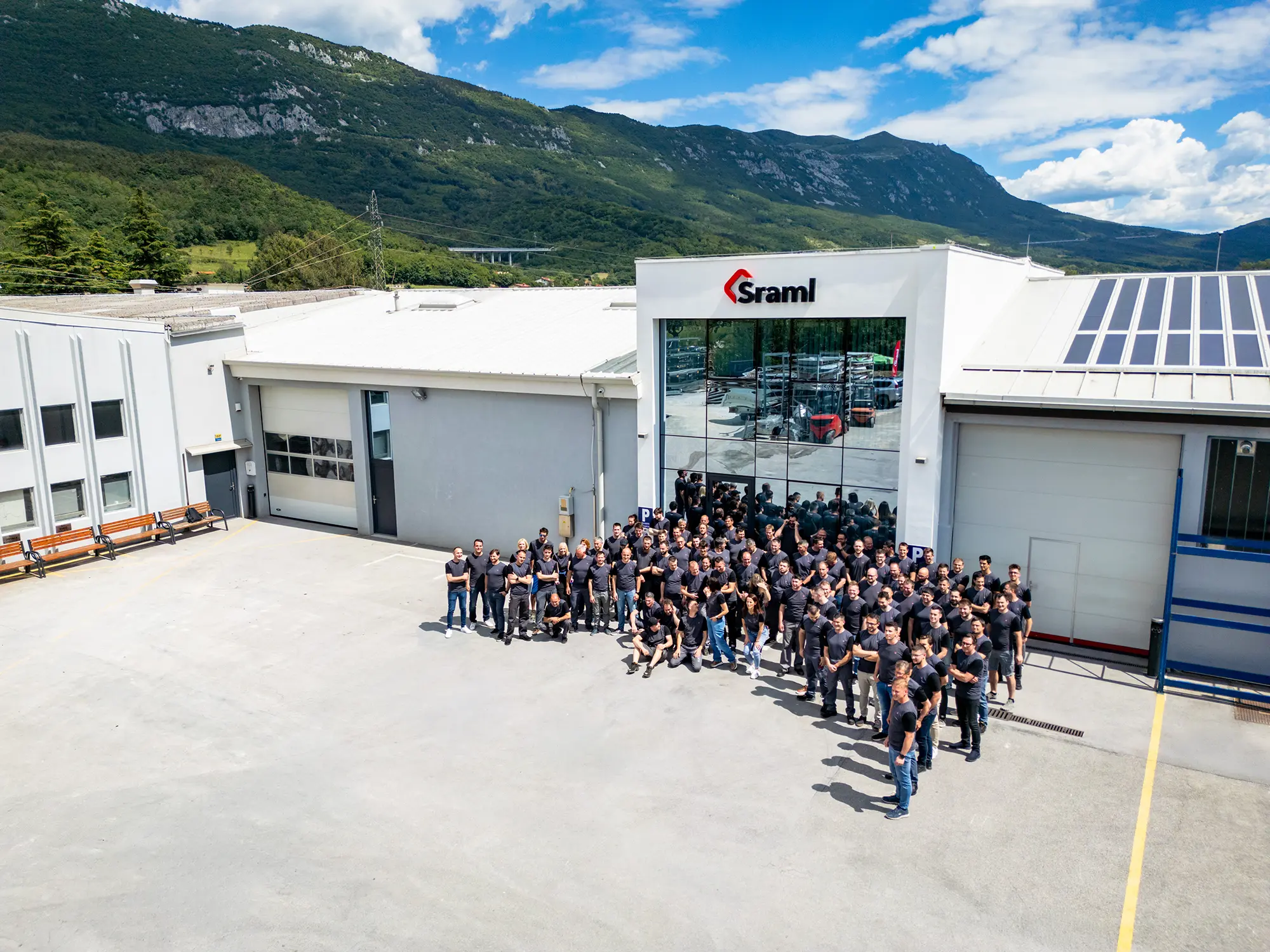 Group of people in dark shirts posing outside a white industrial building with a glass entrance and mountainous backdrop.